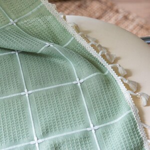 Op de afbeelding: Een saliegroene wafelgeweven deken met een wit rasterpatroon en decoratieve kwastjes langs de rand. De deken is gedrapeerd over een cr&egrave;mekleurig oppervlak, wat de textuur en het ontwerp laat zien. Dit textielartikel is geschikt voor woondecoratie.