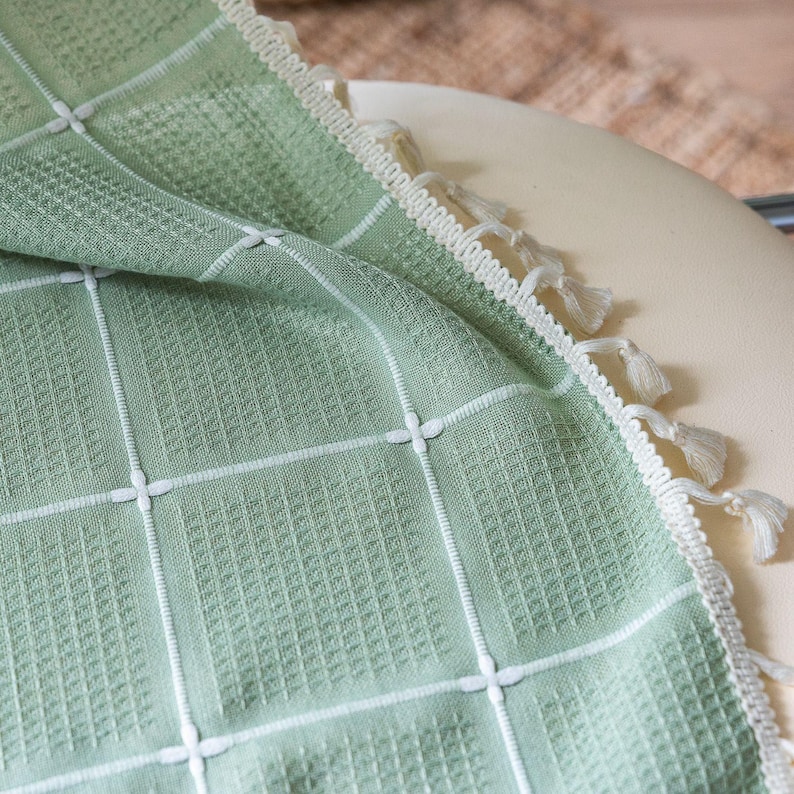 Op de afbeelding: Een saliegroene wafelgeweven deken met een wit rasterpatroon en decoratieve kwastjes langs de rand. De deken is gedrapeerd over een cr&egrave;mekleurig oppervlak, wat de textuur en het ontwerp laat zien. Dit textielartikel is geschikt voor woondecoratie.