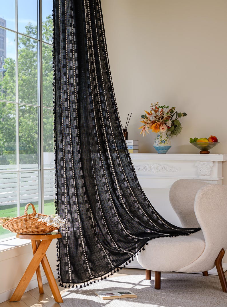 May include: Black sheer curtain with a geometric pattern of squares and vertical stripes. The curtain has a fringe of small black tassels along the bottom edge. The curtain is hanging in front of a window, with a view of a building and trees.