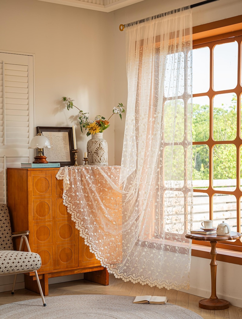 May include: A sheer, off-white curtain panel featuring an intricate, repeating floral vine embroidery pattern and a scalloped lace edge. The lightweight fabric hangs from a rod, partially covering a window with a grid frame and allowing light through.