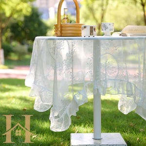 May include: A white lace tablecloth with floral patterns and ruffled edges covers a table. A woven basket with a bottle and fruit sits on the table, along with a small projector and a floral teacup. The table is outdoors on a grassy area.