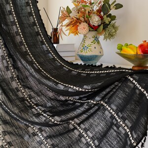 May include: Black curtain with white geometric patterns and tassel trim. The curtain is draped, revealing a glimpse of a vase with flowers and a bowl of fruit. The fabric has a textured appearance.