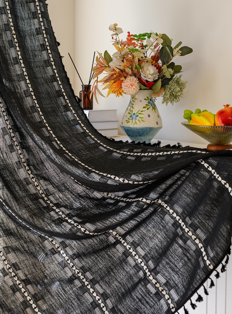 May include: Black curtain with white geometric patterns and tassel trim. The curtain is draped, revealing a glimpse of a vase with flowers and a bowl of fruit. The fabric has a textured appearance.