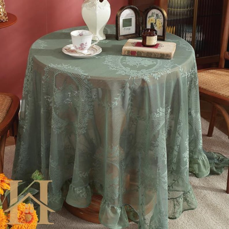 May include: A round, sage green tablecloth with a delicate floral lace pattern. The tablecloth drapes over a wooden table, with a ruffled edge. A teacup and saucer, a vase, and a book are on the table.