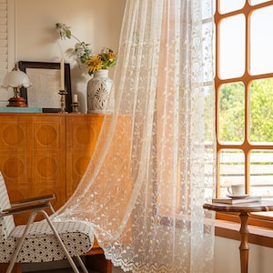 May include: A sheer, off-white lace curtain featuring a delicate, repeating vertical vine and leaf pattern. The fabric is lightweight and semi-transparent, with a scalloped, decorative edge along the bottom hem, hanging in front of a window.