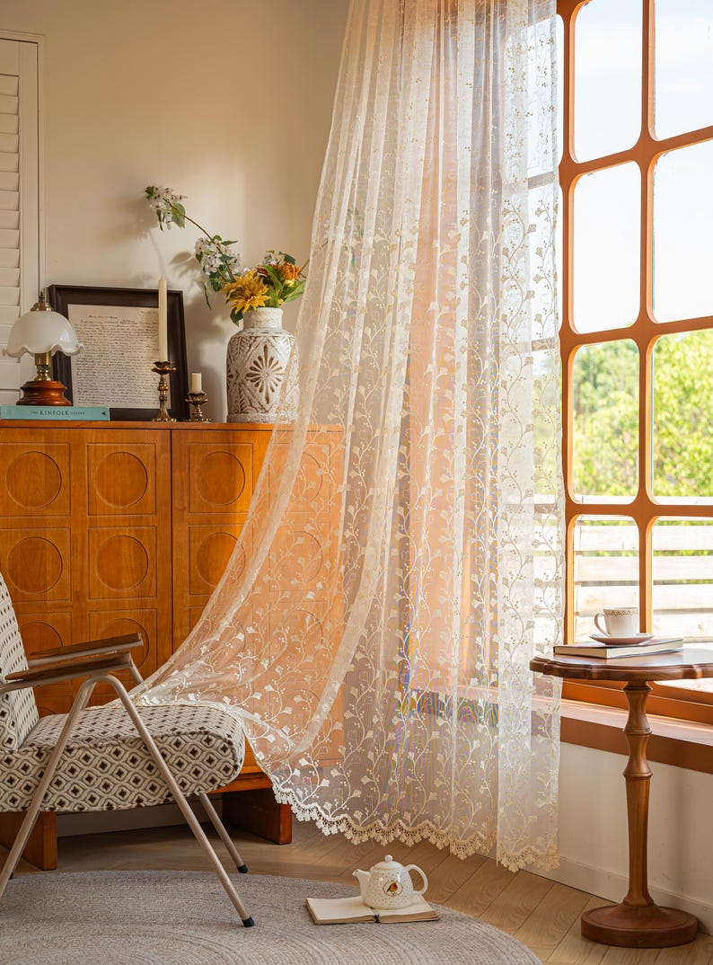 May include: A sheer, off-white lace curtain featuring a delicate, repeating vertical vine and leaf pattern. The fabric is lightweight and semi-transparent, with a scalloped, decorative edge along the bottom hem, hanging in front of a window.