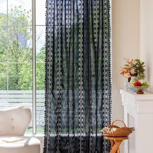 May include: Black sheer curtain with white vertical stripes and square accents. The curtain has a fringe of small black tassels along the edges. The curtain hangs in front of a window, with a view of trees and a white chair.