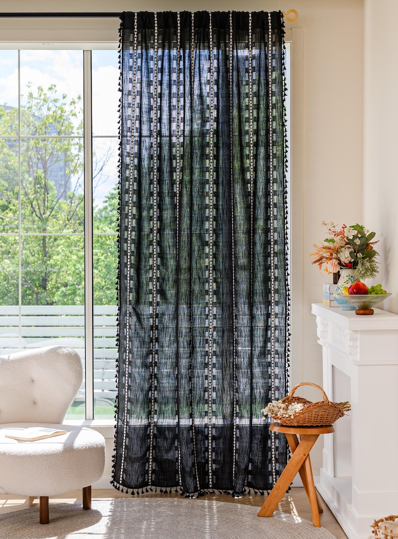May include: Black sheer curtain with white vertical stripes and square accents. The curtain has a fringe of small black tassels along the edges. The curtain hangs in front of a window, with a view of trees and a white chair.