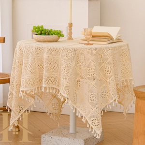 Puede incluir: Un mantel redondo de encaje color crema con un patrón geométrico y flecos. La mesa está decorada con un cuenco de uvas verdes, una vela, una copa y un libro abierto. La mesa está sostenida por un pedestal blanco.