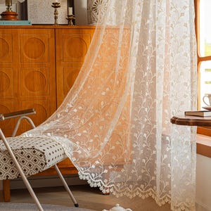 May include: A sheer white curtain panel featuring an intricate embroidered floral vine pattern and a scalloped lace edge. The lightweight fabric hangs vertically, displaying delicate botanical motifs throughout the translucent material.