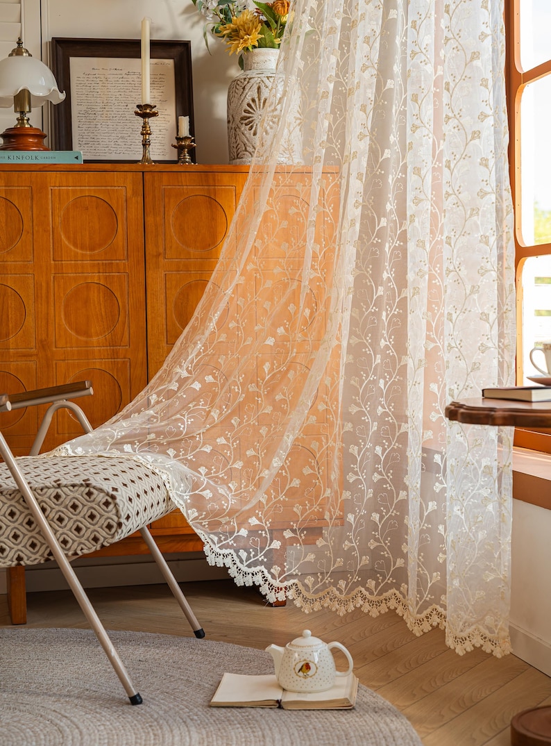 May include: A sheer white curtain panel featuring an intricate embroidered floral vine pattern and a scalloped lace edge. The lightweight fabric hangs vertically, displaying delicate botanical motifs throughout the translucent material.
