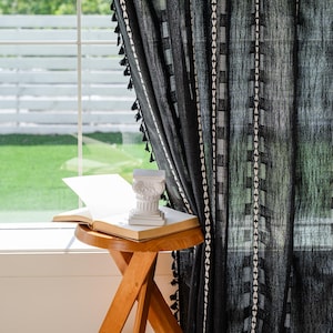 May include: Black sheer curtain with white geometric patterns and black tassels. A small wooden side table holds an open book and a white decorative column. The scene is set near a window with a view of greenery.