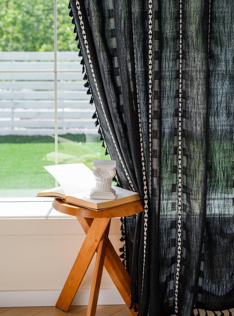 May include: Black sheer curtain with white geometric patterns and black tassels. A small wooden side table holds an open book and a white decorative column. The scene is set near a window with a view of greenery.