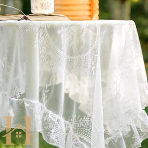 May include: A white lace tablecloth with intricate floral and leaf patterns. The tablecloth has a ruffled edge and is draped over a table. The delicate design adds a touch of elegance to any dining setting.