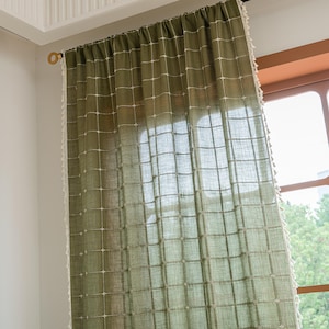 Op de afbeelding: Olijfgroen gordijn met een rasterpatroon en witte rand. Het gordijn hangt aan een goudkleurige stang en bedekt gedeeltelijk een raam. De stof lijkt linnen of een vergelijkbaar gestructureerd materiaal te zijn.