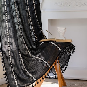 May include: Black curtain with white geometric patterns and tassel trim. The curtain is draped over a wooden stool, with an open book and a decorative column on top. The background includes a fireplace and a shelf with books.