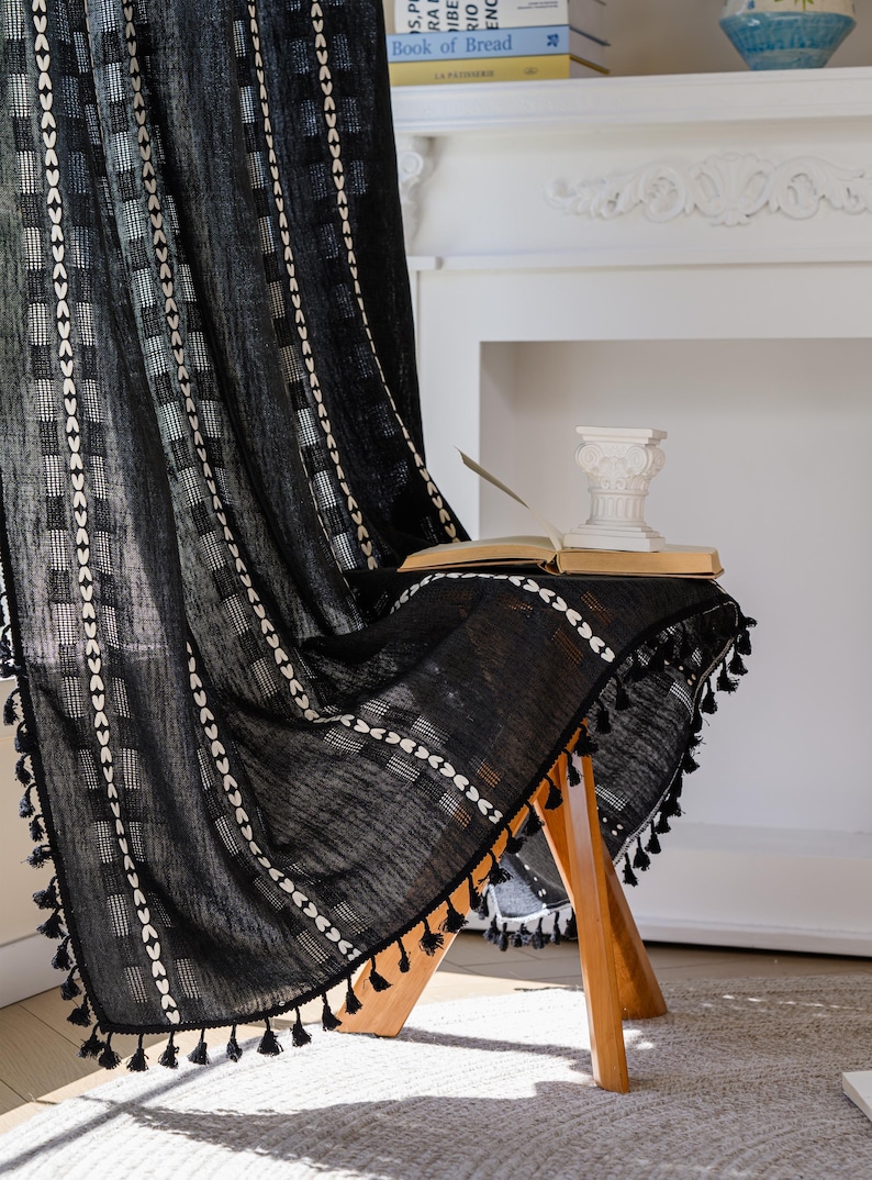 May include: Black curtain with white geometric patterns and tassel trim. The curtain is draped over a wooden stool, with an open book and a decorative column on top. The background includes a fireplace and a shelf with books.