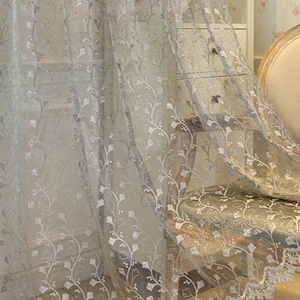May include: A sheer, light gray curtain panel featuring an intricate, embroidered white floral vine pattern that climbs vertically. The bottom edge of the curtain is finished with a scalloped, decorative lace border with detailed stitching.