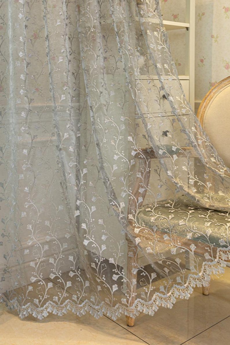 May include: A sheer, light gray curtain panel featuring an intricate, embroidered white floral vine pattern that climbs vertically. The bottom edge of the curtain is finished with a scalloped, decorative lace border with detailed stitching.
