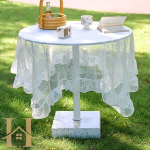 May include: A small, round white table with a white lace tablecloth. The table has a single white leg and a square, stone-like base. A small radio, a cup, and a book are on the table. A basket with fruit sits on the table.