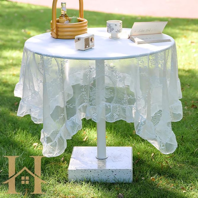 May include: A small, round white table with a white lace tablecloth. The table has a single white leg and a square, stone-like base. A small radio, a cup, and a book are on the table. A basket with fruit sits on the table.