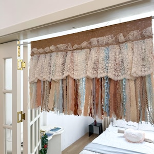 May include: A decorative window valance with a brown burlap top and a layered design. The valance features a white lace trim and hanging ribbons in shades of beige, blue, and brown. The valance is designed to hang above a window or doorway.