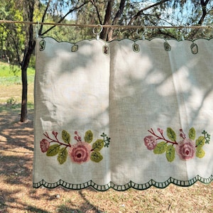 Può includere: Una tenda in lino beige con bordo inferiore smerlato e ricamo verde scuro. Il tessuto è decorato con due motivi floreali composti da rose rosa, foglie verdi e piccole bacche rosse. È appesa a una corda con anelli di metallo.