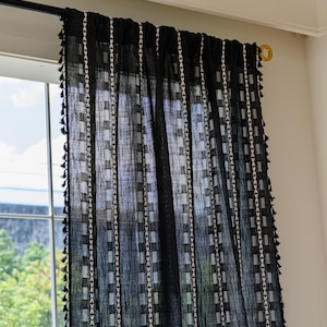 May include: Black sheer curtain with a geometric pattern of white squares and vertical stripes. The curtain has a fringe of small black tassels along the edges. The curtain is hanging in front of a window.
