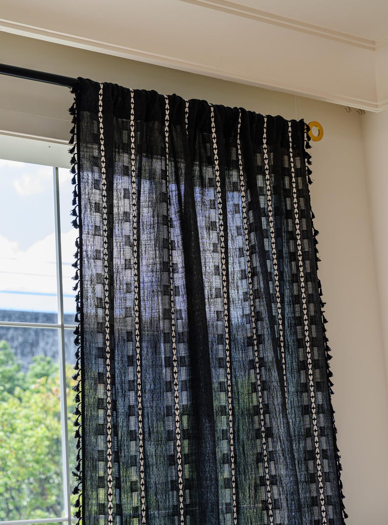 May include: Black sheer curtain with a geometric pattern of white squares and vertical stripes. The curtain has a fringe of small black tassels along the edges. The curtain is hanging in front of a window.