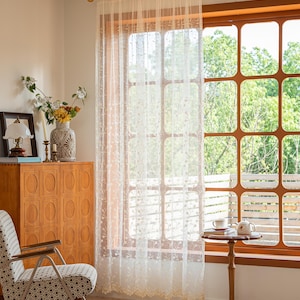 May include: A sheer, off-white curtain panel featuring delicate, embroidered floral vine patterns and a scalloped bottom edge. The lightweight fabric hangs from a dark rod, allowing soft light to filter through while covering a large window.