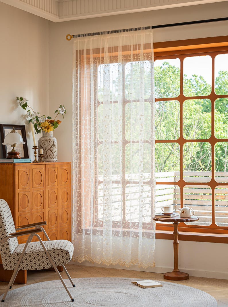 May include: A sheer, off-white curtain panel featuring delicate, embroidered floral vine patterns and a scalloped bottom edge. The lightweight fabric hangs from a dark rod, allowing soft light to filter through while covering a large window.