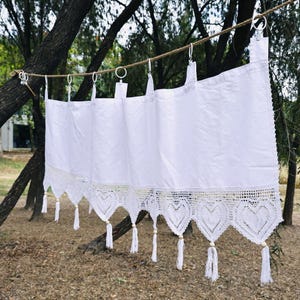 May include: White curtain with a crocheted lace trim featuring heart shapes and tassels. The curtain is hanging from metal rings on a rope, with a natural outdoor background.