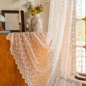 May include: A sheer off-white curtain panel featuring an intricate embroidered floral vine pattern and a scalloped lace edge. The lightweight fabric hangs vertically, displaying a delicate botanical design with small leaves and stems throughout.