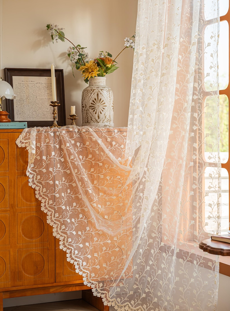 May include: A sheer off-white curtain panel featuring an intricate embroidered floral vine pattern and a scalloped lace edge. The lightweight fabric hangs vertically, displaying a delicate botanical design with small leaves and stems throughout.
