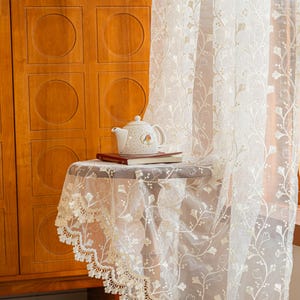 May include: A sheer white curtain featuring an intricate embroidered floral vine pattern and a scalloped lace edge. The fabric is translucent, allowing light to pass through, and the design includes delicate stems and leaves in a repeating vertical arrangement.
