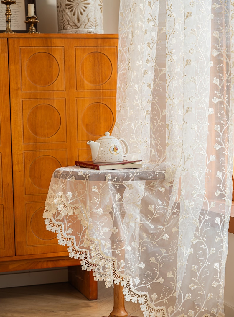 May include: A sheer white curtain featuring an intricate embroidered floral vine pattern and a scalloped lace edge. The fabric is translucent, allowing light to pass through, and the design includes delicate stems and leaves in a repeating vertical arrangement.