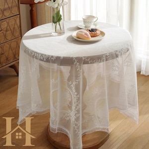 May include: A round, white lace tablecloth draped over a wooden table. The tablecloth features intricate floral patterns and a delicate scalloped edge. A plate with pastries and a teacup sit on top, creating an elegant setting.