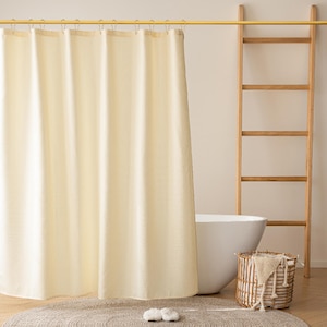 May include: A cream-colored waffle-weave shower curtain hangs from a gold rod. A wooden ladder leans against the wall, and a white bathtub is partially visible. A woven basket with a towel and a round rug complete the bathroom scene.