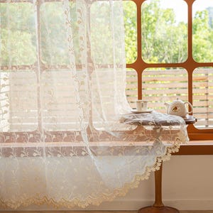 May include: A sheer, off-white lace curtain featuring a delicate, repeating floral vine pattern and a scalloped, embroidered bottom edge. The fabric is lightweight and semi-transparent, allowing light to pass through its intricate, woven design.