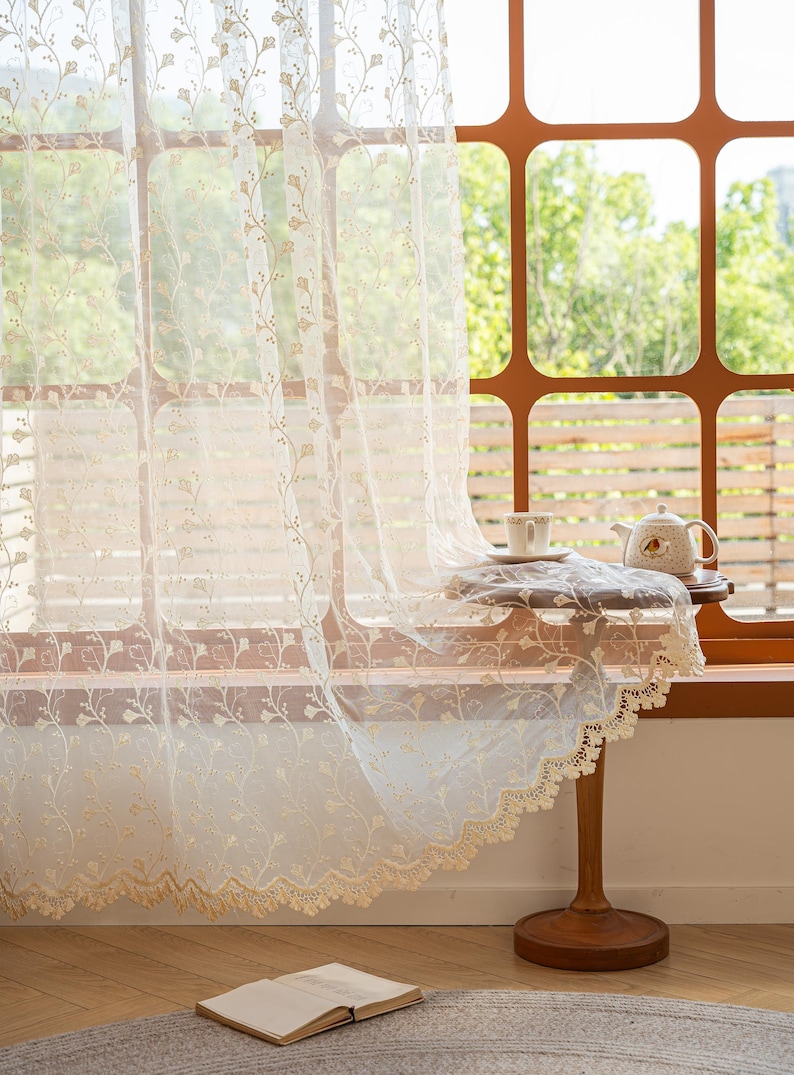 May include: A sheer, off-white lace curtain featuring a delicate, repeating floral vine pattern and a scalloped, embroidered bottom edge. The fabric is lightweight and semi-transparent, allowing light to pass through its intricate, woven design.