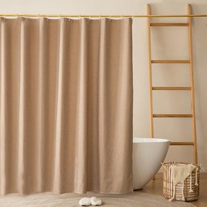 May include: A beige shower curtain hangs from a gold rail, with a wooden ladder and a white bathtub in the background. A woven basket with a towel and a rug with two white slippers complete the bathroom scene.