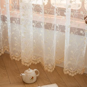 May include: Sheer white curtains featuring delicate embroidered vine patterns with small leaves and a scalloped lace trim along the bottom edge. The fabric is translucent, allowing light to pass through while displaying the intricate floral embroidery.