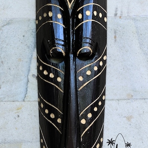 May include: Black wooden mask with gold painted geometric designs. The mask has a stylized face with large eyes and a long nose. The mask is decorated with a pattern of dots and lines.