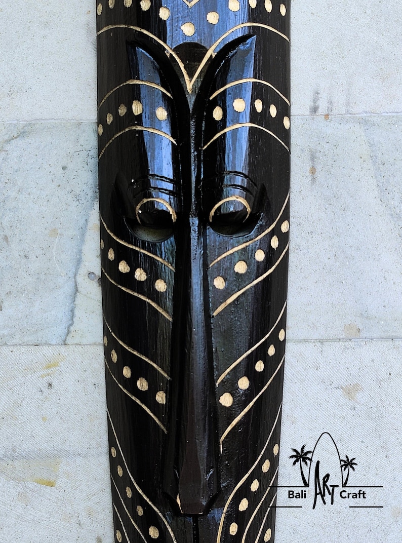 May include: Black wooden mask with gold painted geometric designs. The mask has a stylized face with large eyes and a long nose. The mask is decorated with a pattern of dots and lines.