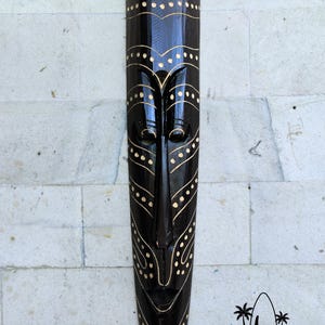 May include: A black wooden mask with gold painted details. The mask has a long, narrow shape with a stylized face and geometric patterns. The mask is hanging on a white tiled wall.