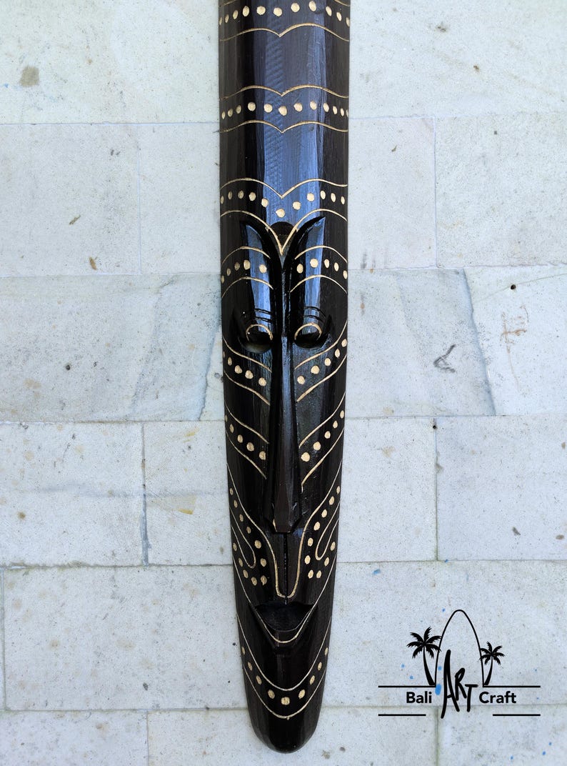 May include: A black wooden mask with gold painted details. The mask has a long, narrow shape with a stylized face and geometric patterns. The mask is hanging on a white tiled wall.