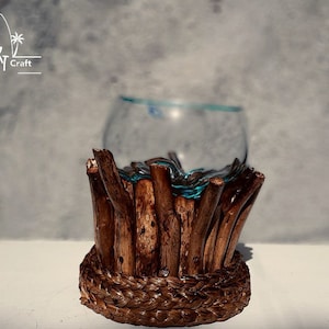 Può includere: Un vaso di vetro trasparente poggia su una base di legno fatta di bastoncini intrecciati e corda. La base è marrone e il vaso è trasparente con una tinta blu.