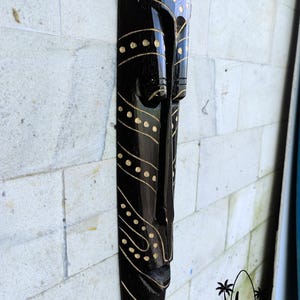 May include: Black wooden mask with gold painted details. The mask has a long, narrow shape and features a stylized human face with a wide mouth and closed eyes. The mask is decorated with a pattern of dots and lines.