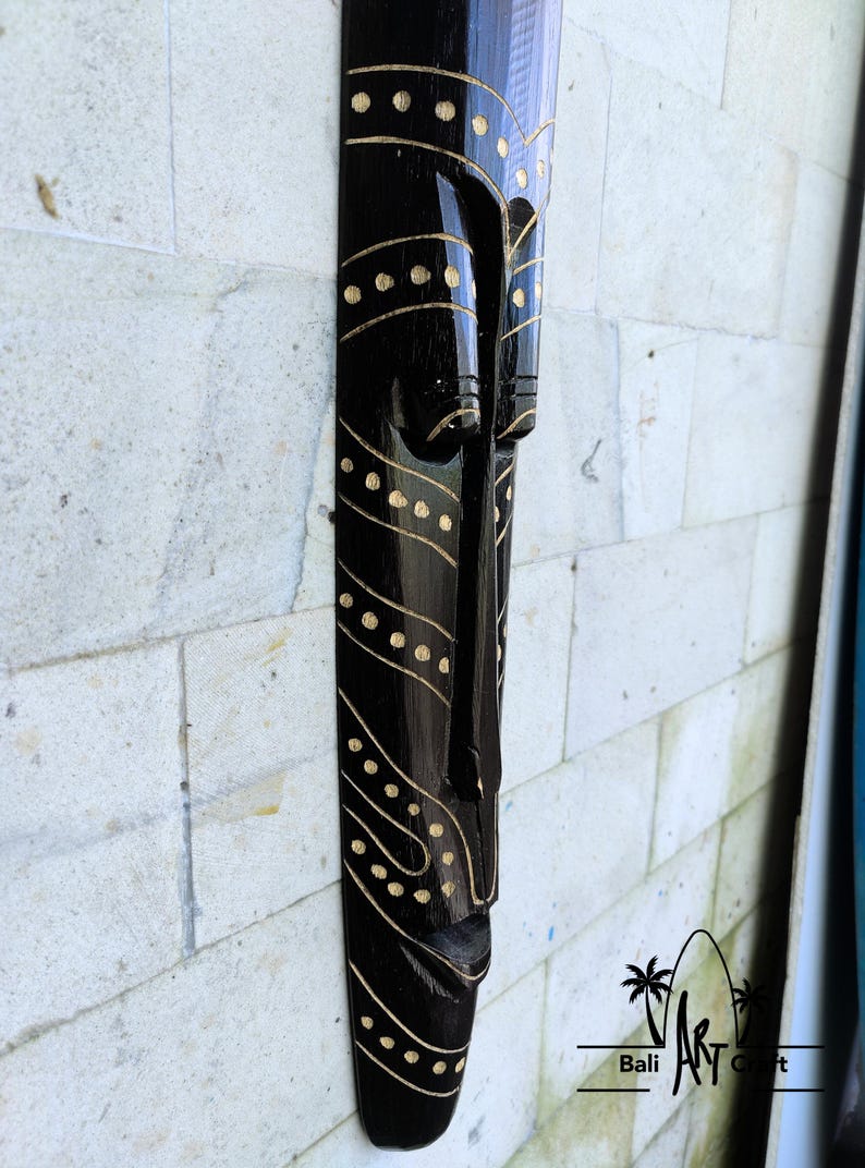 May include: Black wooden mask with gold painted details. The mask has a long, narrow shape and features a stylized human face with a wide mouth and closed eyes. The mask is decorated with a pattern of dots and lines.
