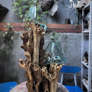 Op de afbeelding: Een decoratief middelpunt met twee heldere glazen vazen op een natuurlijke houten basis. De vazen bevatten water en groen. De houten basis heeft een unieke, getextureerde uitstraling. Het Bali Art Craft-logo is zichtbaar.